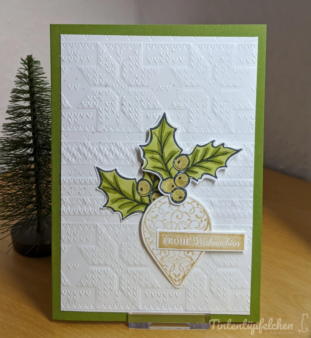 Eine grüne Klappkarte mit einer weißen Prägekarte, die mit einem Muster aus kleinen Dreiecken versehen ist. Darauf sind gestempelte und koloriere Stechpalmenblätter mit Beeren sowie ein goldfarbener Anhänger mit der Aufschrift "Frohe Weihnachten" angebracht.