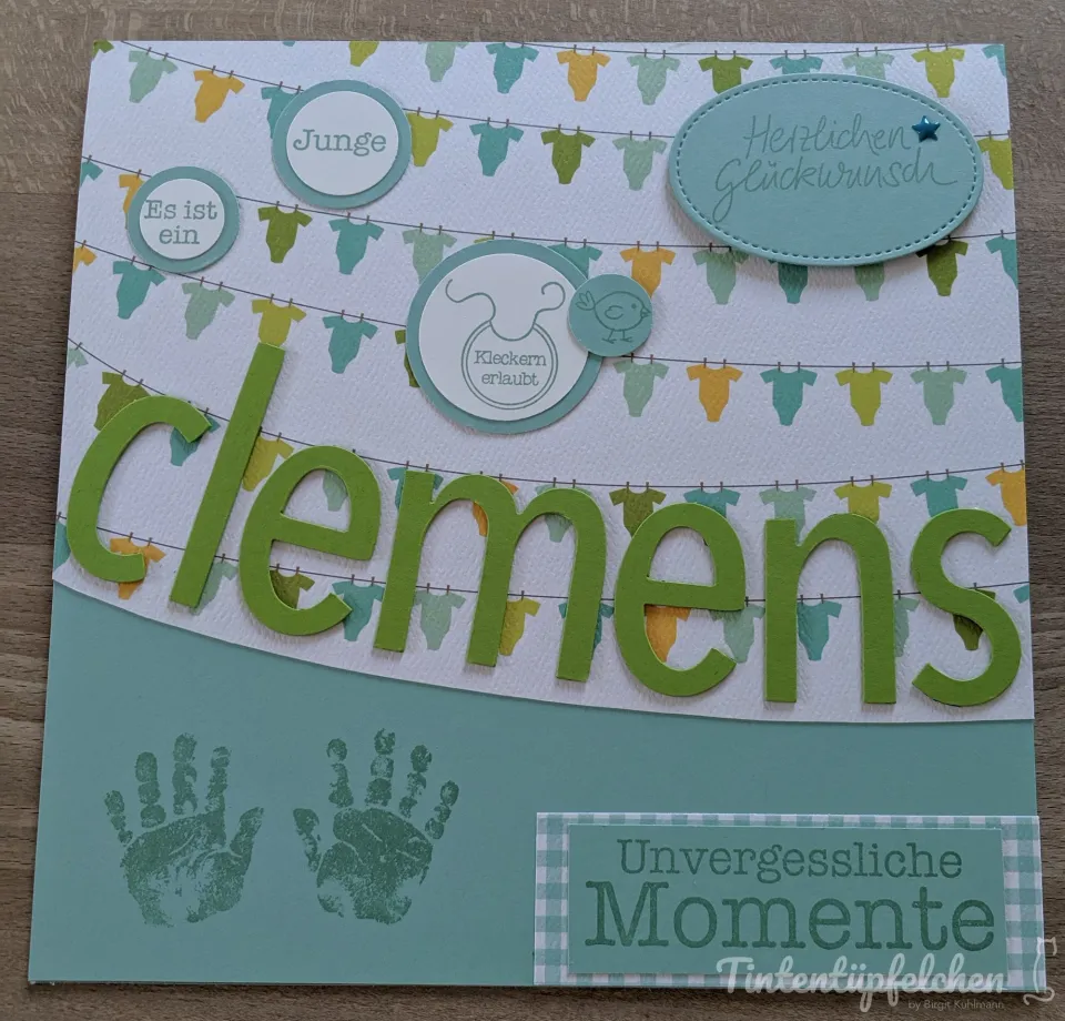Eine Karte zur Geburt mit dem Namen "clemens" in grünen Buchstaben, Schnullern und Babykleidung auf einer Wäscheleine. Darunter sind zwei grüne Handabdrücke und der Spruch "Unvergessliche Momente" zu sehen.