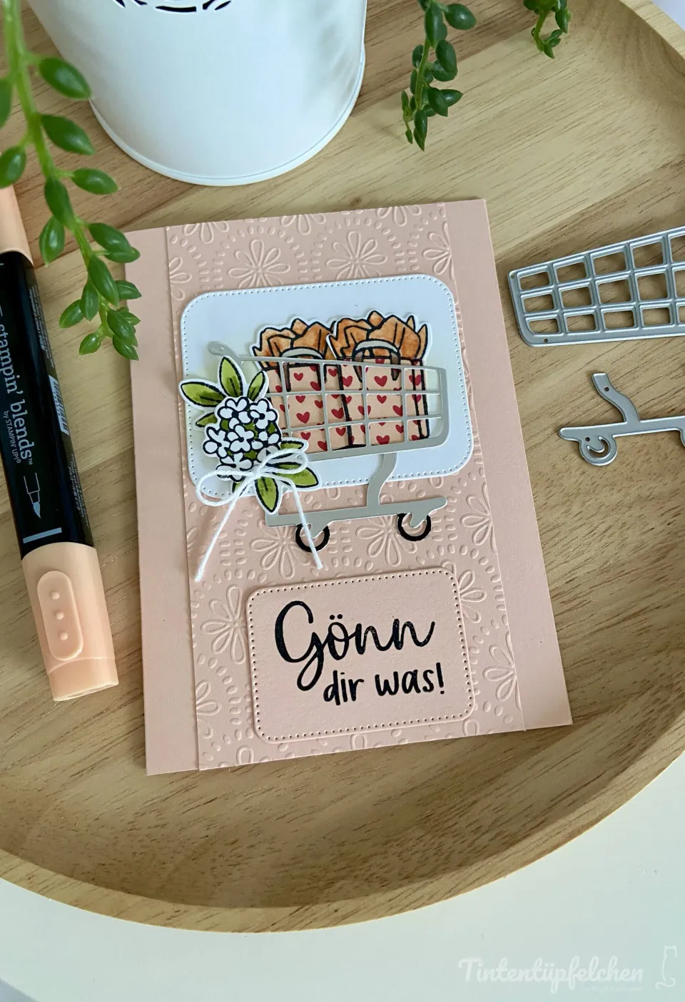 Eine handbastelte Karte mit rosa Hintergrund und eingestampften Blumenmotiven zeigt einen gestanzten Einkaufswagen, gefüllt mit geschnitzelten Taschen und einer kleinen Blumendekoration. Der Text „Gönnt dir was!" ist in schwarzer Tinte auf einem abgesetzten Feld gedruckt, umgeben von Metallschablonen und einem Farbstift.